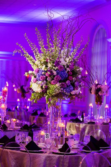 Purple uplighting enveloped the reception space, which featured round tables decorated with towering centerpieces showcasing ivory and purple blooms. Purple Decor Wedding, Purple Wedding Arrangements, Purple Uplighting, Purple Wedding Reception, Purple Flower Arrangements, Purple Reception, Purple Wedding Decorations, Ballroom Reception, Violet Wedding