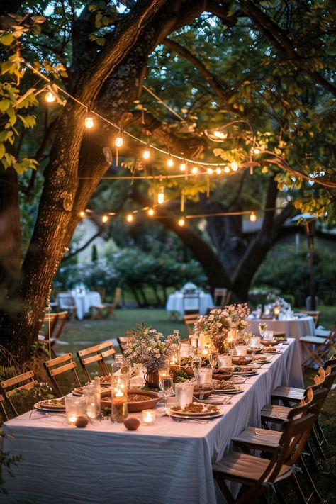 Outdoor table setup with white cloth, candles, and flower centerpieces under string lights hanging from tree branches in a garden during twilight. Garden Celebration Party Ideas, Barbecue Themed Party, Backyard Garden Dinner Party, Backyard Autumn Wedding, Outdoor Garden Party Ideas Decoration, Elegant Backyard Dinner Party, Summer Garden Dinner Party, Engagement Party Garden, Birthday Decor Outdoor