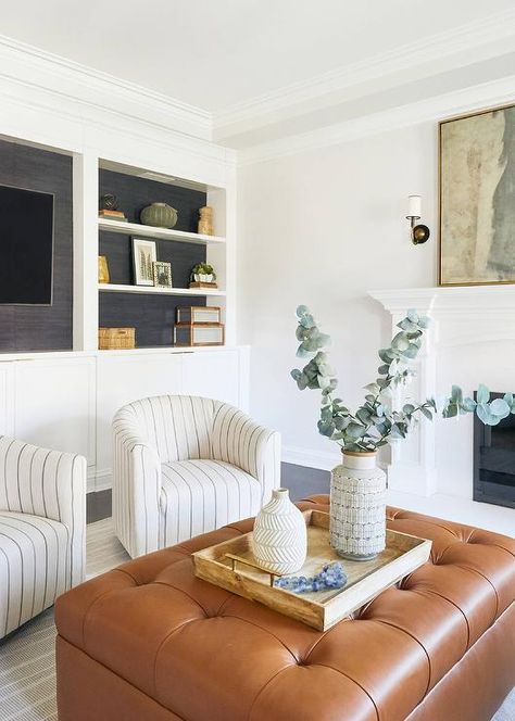 Light, airy and styled to perfection- this living room is furnished with ivory and gray stripe swivel chairs facing a brown leather ottoman atop a gray and white stripe area rug. Leather Ottoman Styling, Brown Ottoman Living Room, Leather Ottoman Decor, Leather Ottoman Living Room, Leather Ottomans Living Room, Hoboken Apartment, Brown And Blue Living Room, Brown Leather Ottoman, Charleston House