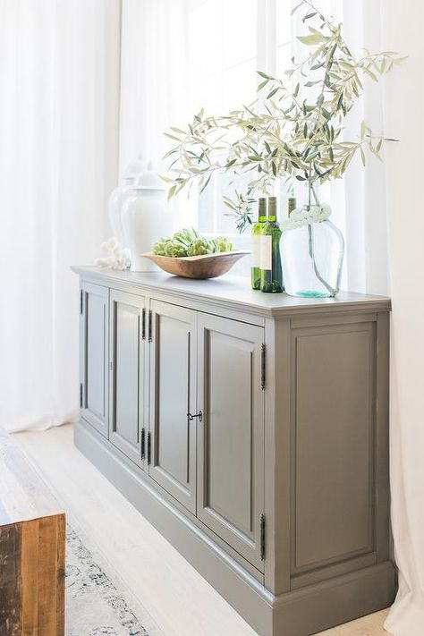 Adding a dark gray buffet cabinet in a dining room with white furnishings and lots of natural light is the perfect way to balance the space. Sideboard Decor In Front Of Window, Family Room Sideboard, Gray Sideboard Buffet, Kitchen Buffet Cabinet Traditional, Transitional Buffet Cabinet, Dining Room Decor Sideboard, Buffet Cabinet In Front Of Window, Grey Buffet Table, Side Buffet Decor Dining Rooms