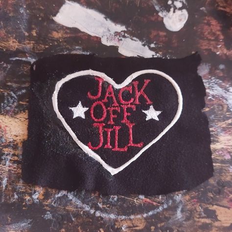 An unevenly cut, black, rectangle, fabric patch is on a paint-stained background. On the patch is a white heart, inside the heart, written in red, are the words "Jack Off Jill" in a vertical column. On either side of the word "Off" are white stars. Jack Off Jill Logo, Alternative Accessories Diy, Punk Shirts Diy, Punk Pins And Patches, Punk Patch Stencil, Punk Band Patches, Band Patches Diy, Punk Diy Ideas, Jack Off Jill Band