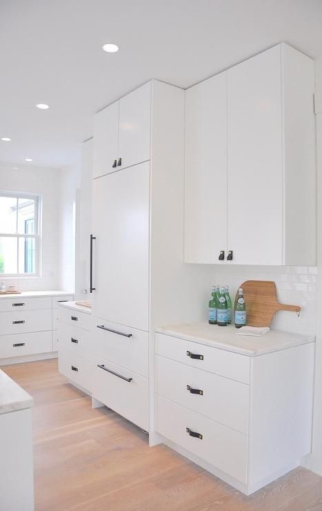 Black leather tab pulls beautifully contrast white flat front cabinets finished with a honed white marble countertop mounted against white linear backsplash tiles, as upper cabinets flank a camouflaged refrigerator, recessed and fitted with matte black handles. White Flat Kitchen Cabinet Doors, White Fridge Black Handles, White Cabnits With Black Handles, White Cabinets Black Hardware, Townhome Kitchen, White Kitchen Cabinet Handles, White Cabinet Hardware, Slab Kitchen Cabinets, Flat Front Cabinets