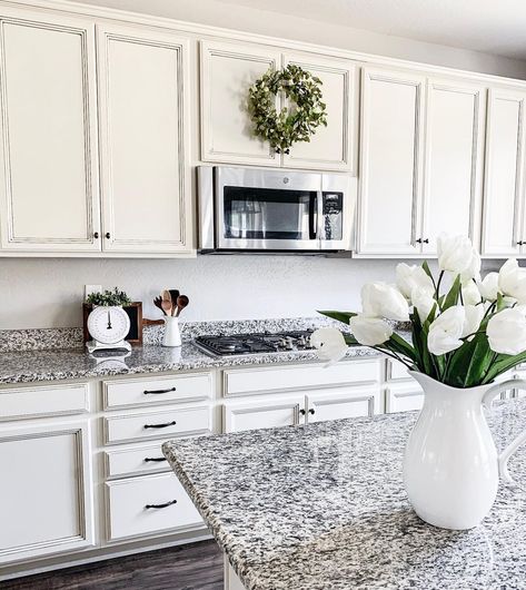 This kitchen features black and white backsplash with granite counters. White cabinets with black hardware encase stainless steel microwave tucked above gas range. Porcelain pitcher with an array of white flowers is set upon countertop. Granite Countertops With White Cabinets Grey Backsplash Ideas, White Cabinets Black And White Granite, Granite Backsplash With Tile Above, Black And White Kitchen Granite, White And Black Granite Countertops, Black And White Granite Kitchen, Black And White Granite, Black And White Granite Countertops, Granite Countertops With White Cabinets