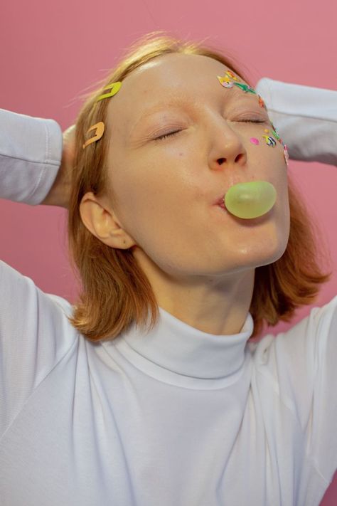 woman blows yellow bubble gum from her mouth. her eyes are closed and she wears a white turtle neck top. she has bright hair clips in her hair and stickers around her left eye. there is a pink backdrop behind her. her arms are behind her short red hair. Chewing Gum Jawline, Girl Chewing Gum Aesthetic, Stickers On Face, Center Fresh Chewing Gum, Chewing Gum Meme Funny, Extra Chewing Gum, Free Person, Fun Portraits, How To Remove Warts