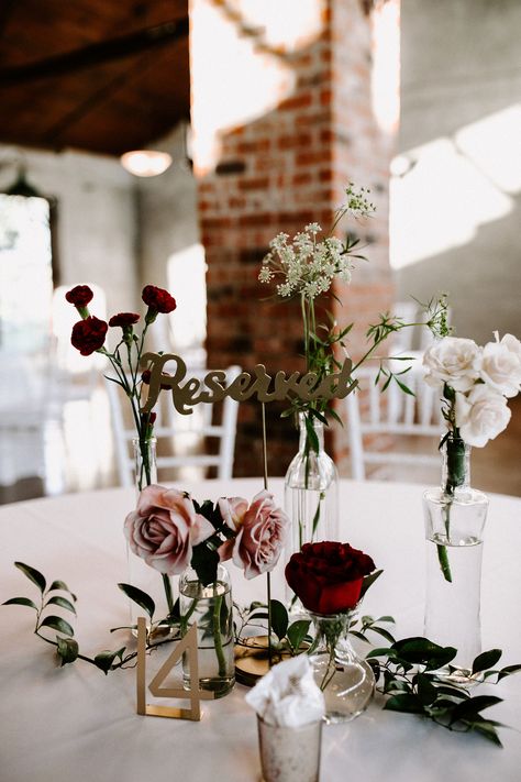 Wedding table setting with roses and other flowers in bud vases and table number. Bud Vases Wedding Centerpiece Winter, Burgundy Bud Vase Centerpiece, Black And Burgundy Centerpieces, Burgundy Bud Vases, Moody Bud Vases, Single Flower Centerpiece, Wedding Centerpiece Burgundy, Vase Wedding Centerpieces, Priston Mill