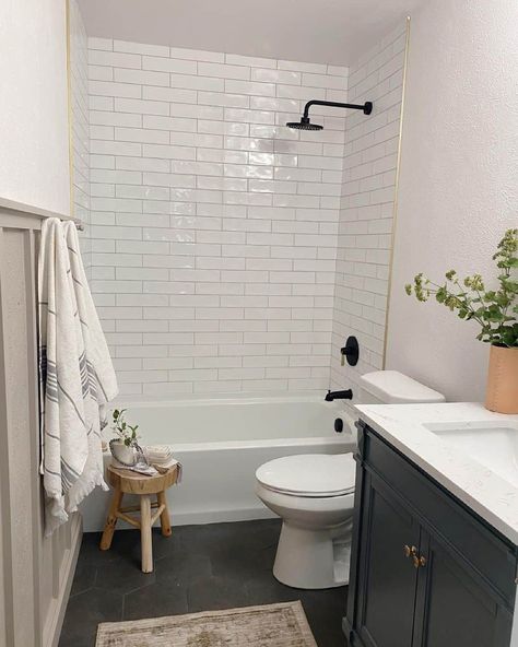 This gray and white farmhouse bathroom features a white subway-tiled shower and bathtub with gray flooring and dark painted cabinetry. Next to the tub rests a three-legged stool accented with greenery. White Farmhouse Bathroom, Dark Tile Bathroom, Bathroom Remodel Plans, Gray Flooring, White Subway Tile Bathroom, Shower And Bathtub, Farmhouse Gray, Condo Bathroom, Subway Tile Showers