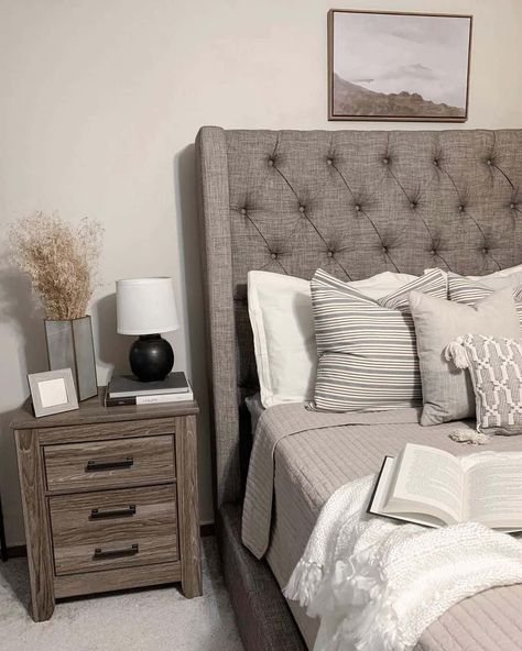 Relax in this beige and grey bedroom featuring a grey tufted bed outfitted with white and grey bedding. The bed is styled with wood frame artwork mounted above the headboard. A stained wood night table perched next to the bed is used to display beige, white, and black décor. White And Grey Bedding, Grey Farmhouse Bedroom, Grey Tufted Bed, Grey Bed Frame Bedroom Ideas, Tufted Beds, Cream And Grey Bedroom, Grey Tufted Headboard, Beige And Grey Bedroom, Grey Headboard Bedroom Ideas