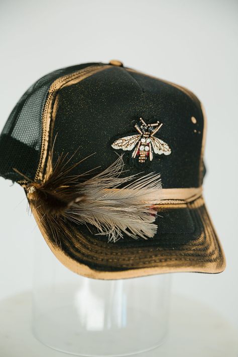 Look sharp and show off your style with this GOLDEN BEE BLACK TRUCKER HAT. This unique hat features a black trucker hat with gold accents, a gold bee patch, and a black and white feather combo that is sure to turn heads! Rock this hat and look cool and stylish - no bee-lieving required! This is a made-to-order item. All customized orders are currently shipping within 14 business days. To receive item quicker, expedited shipping is available at checkout. Bridal Trucker Hats, Cool Hat Designs, Women’s Trucker Hats, Trucker Hats With Patches Diy, Diy Trucker Hat Ideas, Cute Trucker Hats, Painted Trucker Hats, Custom Hat Ideas, Trendy Hats For Women