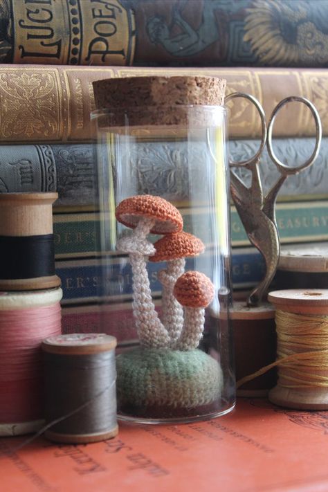 A collection of items placed in front of a stack of vintage books, including spools of thread, a pair of vintage bird scissors, and a glass jar filled with three orange capped micro crochet mushrooms with white stems sitting on a green mound of moss. Crochet Mushroom Things, Crochet Home Goods, Dark Cottagecore Crochet, Goblincore Crochet Ideas, Crochet Terrarium, Crochet Display Ideas, Cottagecore Amigurumi, Stash Jar Crochet Pattern, Crochet Mushroom Jar