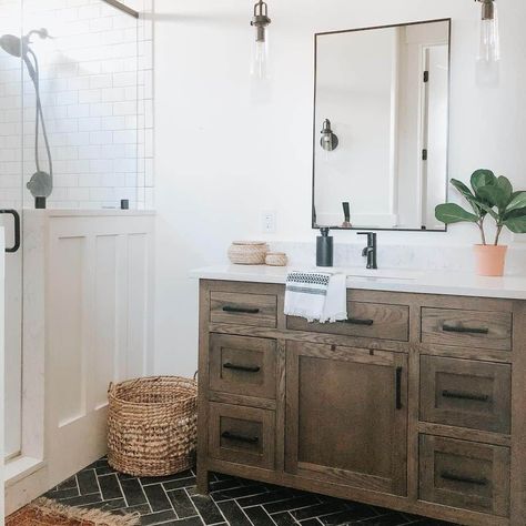 A green plant sits on the white bathroom counter of a rustic wood vanity. A glass door reveals a white subway tile walk-in shower wall, and a brown mat is laid on the black herringbone tile floor...   Image: hestershomestead Lauren Diy, Home Depot Vanity, Black Sconces, Neutral Bathroom, Black Faucet, Black Wall Mirror, Vanity Ideas, Bathroom Wall Sconces, Wood Vanity