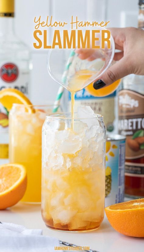 A yellow hammer slammer is being poured into a tall glass.  Several bottles and fresh cut oranges rest in the background. Yellow Alcoholic Punch, Yellow Hammer Slammer, Yellow Hammer Drink Recipe, Yellow Mixed Drinks, Yellow Hammer Slammer Drink, Yellow And Red Cocktails, Yellow Party Drinks, Yellow Alcoholic Drinks For A Party, Yellow Drinks For Party