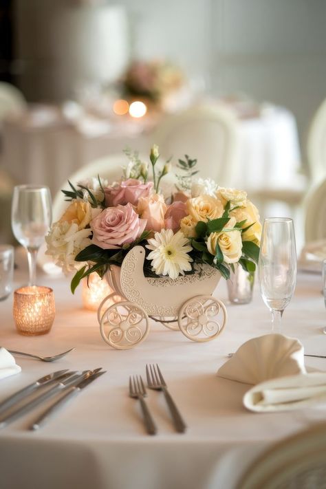 A decorative table centerpiece ideal for baby shower centerpiece ideas, featuring a small white carriage filled with pastel flowers like roses and gerberas. The table is elegantly set with glassware and cutlery, surrounded by softly lit candles on a pristine white tablecloth. Baby Shower Centerpiece Ideas, Baby Shower Elegante, Babyshower Decor, Diy Baby Shower Centerpieces, Girl Baby Shower Centerpieces, Baby Shower Table Centerpieces, Shower Flowers, Storybook Baby Shower, Baby Shower Table Decorations