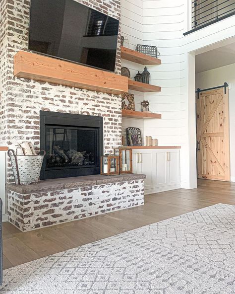 Two story limewash brick fireplace is finished with a gray stone hearth and light stained wood mantel. A flat panel TV is mounted above the mantel in this white shiplap living room. Floating Shelves Tall Ceiling, 2 Story Built Ins, Black White And Wood Fireplace, Brick Fireplace Floating Shelves, 2 Story Shiplap Fireplace Wall, Floating Shelves High Ceiling, Two Story Brick Fireplace, 2 Story Fireplace Ideas, Wash Brick Fireplace