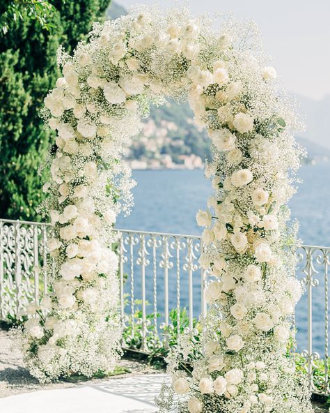 The majestic all-white wedding arch blends seamlessly with this stunning location! White gypsophila, roses and hydrangeas All White Arch Wedding, All White Flower Arch, White Rose Ceremony Arch, White Roses Arch Wedding, Outdoor Wedding Entrance Decor, White Flower Arch Wedding Outdoor, Small Outdoor Ceremony, Wedding Aisle White Flowers, All White Wedding Arch