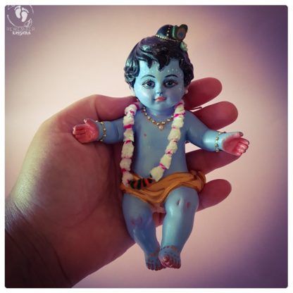 Baby Krishna deity being held in hand against a purple pink backdrop. Baby Krishna figurine is wearing a Pavitra silken thread garland and has a peacock feather in his hair tucked into a little bun. The figure of Krishna statue has his arms out welcoming a hug or bestowing blessings on all. His chubby toes are gently kicking and His face has a shining mellow smile and tilak decorates his forehead. His chest and neck are adorned with golden jewels. Krishna Holding Hand, Small Krishna, Krishna Doll, Krishna Artwork, Krishna Murti, Krishna Birthday, Janmashtami Celebration, Krishna Drawing, Little Krishna