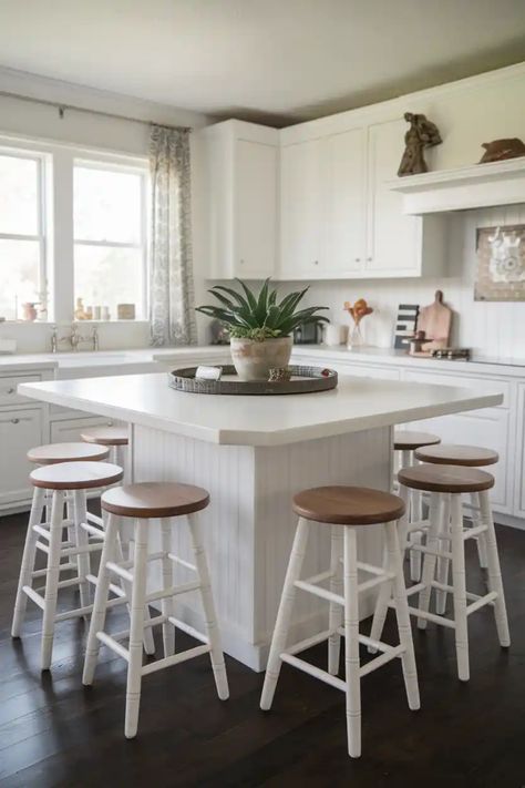 White kitchen with a central island, surrounded by wooden stools, and a potted plant centerpiece. Kitchen Island Tops, Island Stools, White Kitchen Island, Saddle Stools, Stools For Kitchen Island, Bar Stools With Backs, Hillsdale Furniture, Upholstered Stool, Farmhouse Style Kitchen