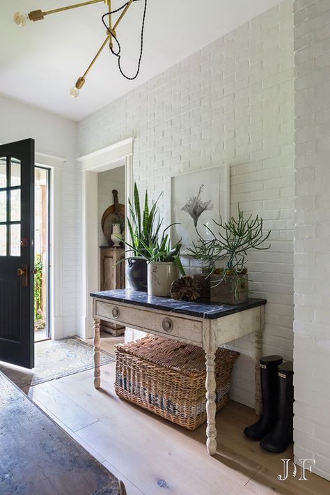 Black front door leads to distressed side table filled with various greenery. The table sits against a white brick wall over a large wicker basket. White Brick Wall Dining Room, White Brick Walls Interior, Painted White Brick Interior, White Brick House Interior, White Brick Entryway, Brick Wall Foyer, White Painted Brick Interior, Brick Wall Lounge, White Brick Wall Decor