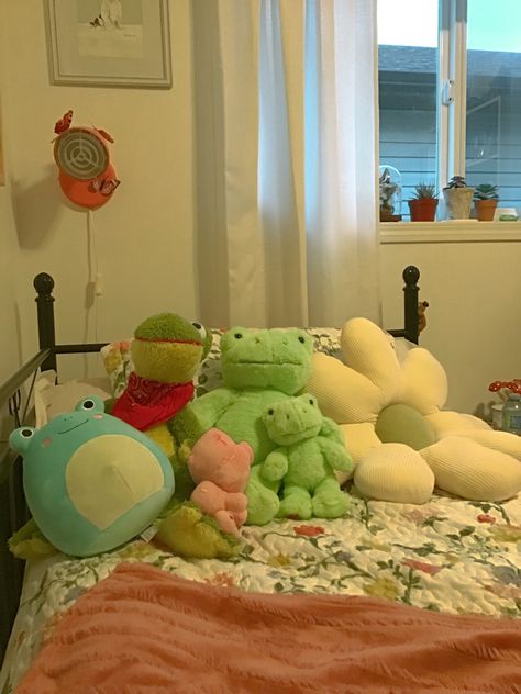 This is a picture of a room. It mostly shows the bed. On the bed is a floral quilt. There is a soft rose coloured throw blanket too. There are also stuffed animals, 4 frogs and a minecraft pig. There is a pillow shaped as a flower that is cream coloured with a green centre. The bed has a black metal frame. Above the bed on the wall is a wall lamp that is coral in colour. There are fake butterflies on it. Above the light is a picture frame with a dog painting in it. There’s a window Stuffies Aesthetic Room, Frog Bedroom Aesthetic, Minecraft Room Decor Aesthetic, Kawaii Cottagecore Bedroom, Buildabear Aesthetic, Frog Bedroom Ideas, Frog Themed Bedroom, Kidcore House, Frog Room Decor