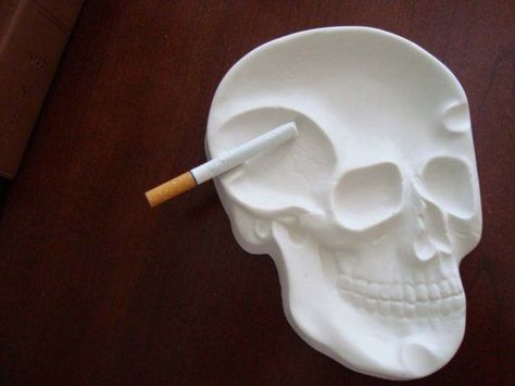 a cigarette sits along the side of a white ashtray. The ashtray is made out of clay, and is shaped like a 2-dimensional human skull. Air Dry Clay Ashtray, Skull Ashtray, Diy Skeleton, Skull Wedding Ring, Skull Engagement Ring, Skull Wedding, Kitchen Counter Top, Skeleton Skull, Ceramic Bottle