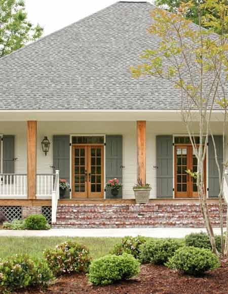 Design by Krista Lewis, K. Lewis Design.  Photography by Nancy Nolan for At Home in Arkansas. Acadian cottage exterior Brick Southern Home Exterior, White House With Natural Wood Shutters, Olive Green And Cream House Exterior, White House With Stained Shutters, Covered Porch Paint Colors, Exterior Oval Window, White And Green Home Exterior, Trendy Shutter Colors, White Cottage With Shutters