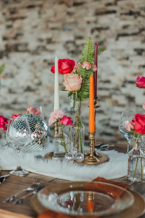 We had so much fun styling this boho disco themed shoot! We used vintage elements in combination with classic boho decor to make a super fun and unique table. These long farm tables are great for styling unique centerpieces. For ours, we used a combination of vintage candlestick holders, bud vases filled with assorted pink flowers, shiny disco balls, and went with a fun fur throw as a table runner. Sparkly and metallic gold chargers looked just right with a warm brown napkin. Shot by @kaboovang Disco Ball Wedding Tables, Bud Vases With Pink Flowers, Fun Flower Vases, Pink Bud Vases Wedding, Centerpieces With Disco Balls, Disco Centerpieces Wedding, Disco Wedding Flowers, Disco Ball Wedding Table Decor, Disco Wedding Table Setting