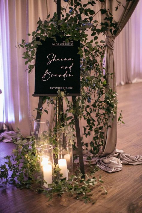 Wedding welcome sign with greenery vines and hurricane candles Wedding Vines Decor, Wedding Welcome Sign Candles, English Ivy Wedding Decor, Green Vines Wedding Decor, Welcome Sign Wedding Greenery, Flower Display Wedding, Wedding Welcome Sign With Candles, Wedding Arch With Candles, Welcome Sign With Candles