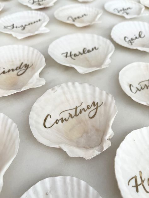 Scallop shells featuring gold calligraphy by Nob Hill Jane are a unique place card idea for your coastal or beach themed wedding. Beach Theme Place Settings, Beach Wedding Table Assignments, Oyster Shell Wedding Place Cards, Seashell Place Cards, Shell Place Cards Wedding, Seashell Name Cards, Coastal Place Settings, Coastal Themed Wedding, Shell Name Cards Wedding