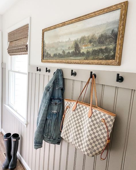 A horizontal painting adds vintage character to a white wall, while the bronze frame infuses a big personality. Modern hooks are installed on beige wainscoting trim to offer contrast and create the perfect place to hang possessions after a long day. Beige beadboard extends across the bottom of the walls, highlighted by white trim. Beige Wainscoting, Rustic Wainscoting Ideas, Horizontal Beadboard Walls, Painted Beadboard Walls, Beadboard Mudroom, Gray Beadboard, Wallpaper And Beadboard, Rustic Wainscoting, Beadboard Trim