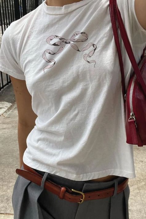 A cropped photo shows a girl from the waist to just below her neck, wearing a white tee shirt with a light pink bow illustration, paired with gray slacks, and a brown leather belt, and cherry red handbag Coquette Graphic Tee, Graphic Baby Tee Outfit, Baby Tees Outfit, Baby Tees Aesthetic, Cute Baby Tees, Baby Band Tees, Baby Tee Aesthetic, Aesthetic Ribbon, Graphic Tee Aesthetic