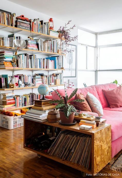 There is a rectangular oak end table in the foreground, with books on the upper and lower shelves and potted plants on top. Next to it there is a cotton candy pink couch with a few white and blush throw pillows. Behind the couch there is a large white wall with five long natural wood bookshelves. There are hundreds of books stacked on top of and next to each other on these shelves. There is a large window in the back window letting in tons of natural light. Colorful Eclectic Living Room, Room Size Rugs, Behind Couch, Boho Interior Design, Eclectic Living Room, White Living Room, Boho Interior, Furniture Arrangement, Home Library