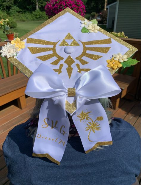 A white graduation cap with a sparkly gold boarder. The hat has yellow and white flowers as well as a sparkling gold triforce symbol. There is a white bow fixed to the bottom corner with the initials SVG  and the words “Drexel 24” on one tail of the bow. The other tail has an image of a silent princess in the same sparkling gold as the rest of the hats detail. Splatoon Graduation Cap, Grad Cap Studio Ghibli, Jjba Graduation Cap, Genshin Graduation Cap, Graduation Cap Designs Coraline, Legend Of Zelda Graduation Cap, Video Game Grad Cap, Hxh Graduation Cap, Undertale Graduation Cap