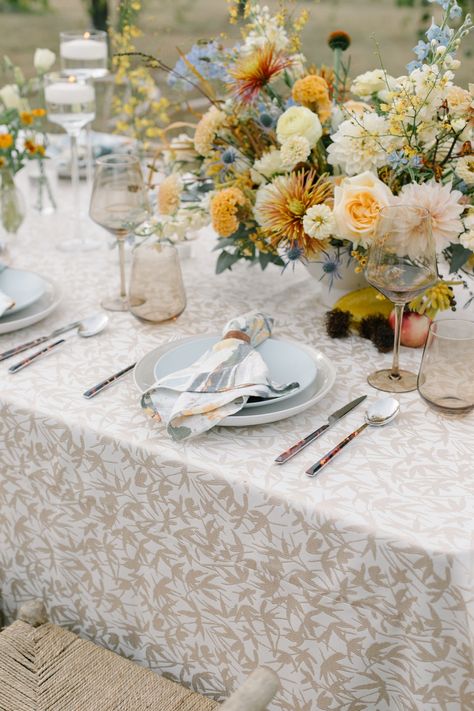 A wildflower floral arrangement and colorful wine glasses atop a neutral patterned linen with wicker chairs and a colorful napkin. Stradley Davidson's Ascot Garden was used for the napkin and Fitz Almond for the table linen. Patterned Wedding Table Linens, Colorful Wedding Linens, Patterned Tablecloth Wedding, Floral Tablecloth Wedding, Wildflower Table Setting, White Wedding Table Setting, Wedding Drinkware, Vintage Table Setting, Reception Table Settings