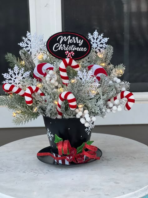 Celebrate the joy and wonder of the winter season with this enchanting snowman hat centerpiece. This unique piece is the epitome of holiday cheer, featuring a charming snowman hat surrounded by festive candy canes, twinkling lights, and lush winter pines. Whether it's the centerpiece of your holiday table or a cheerful addition to your mantel, this delightful arrangement is sure to bring smiles and a cozy, festive atmosphere to your home. Details: *Composition includes a decorative snowman hat, artificial candy canes, LED string lights, and faux winter pine branches *Battery-operated lights for easy and safe use (batteries not included) *Perfect for holiday tablescapes, mantel decor, or as a festive accent piece Mason Jar Winter Centerpieces, Snowman Table Setting, Snowman Table Decorations Centerpieces, Easy Christmas Centerpieces For Table, Snowman Centerpiece Ideas, Christmas Centerpieces Diy Table, Snowman Hat Centerpiece, Candy Cane Centerpiece, Diy Christmas Hats