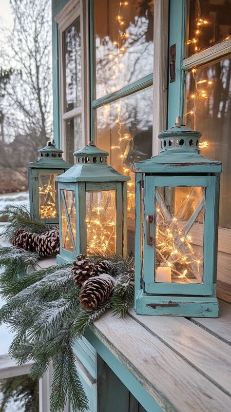 Rustic lanterns with fairy lights on a wooden windowsill, surrounded by evergreen branches and pine cones in muted teal and Wooden Windowsill, Woodsy Farmhouse, Decorating With Lanterns, Candle Centerpieces Elegant, Rustic Candle Centerpieces, Rustic Christmas Decor Ideas, Lantern Aesthetic, Muted Teal, Birch Candles