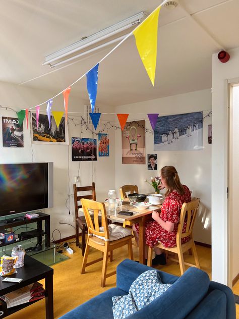 You see a girl in her early twenties sitting at the dinner table in a student house. The girl is wearing a red dress, and it is golden hour so the sun is shining through the window creating a rainbow effect on the wall. The wall is decorated with a bunch of random posters including one of Harry Styles and the women soccer team. The room also includes a yellow floor, a blue couch, and birthday decorations. Student House Living Room, Flat Share Aesthetic, Girls Uni House Aesthetic, Student House Aesthetic, Uni Living Room, Simple Living Room Ideas Apartment, Student House Decor, Uni House Decor, Student Living Room