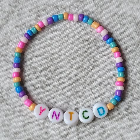 A friendship bracelet with the letters Y N T C D in pink, purple, orange, green and blue letters respectively. The seed beads that surround the letters alternate in a pastel orange, green, blue, purpke, light pink and dark pink pattern. Taylor Swift Lover Era Bracelets, Flat Bead Bracelet Ideas Taylor Swift, Clay Beads Eras Tour Bracelets, Taylor Swift Eras Colours, Yntcd Taylor Swift Bracelet, Lover Era Bracelet, Yoyok Friendship Bracelet Taylor Swift, Yntcd Taylor Swift, Swifty Bracelet