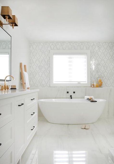 Freestanding oval tub under a window framed by white and gray mosaic wall tiles. A leaning towel ladder is positioned next to a white double vanity complemented with oil rubbed bronze pulls and a white quartz countertop fitted with his and her sinks complemented with brushed gold faucets. The faucets are fixed beneath a black framed mirrors lit by brass 2 light sconces. Image source - Casa Perfecta Bath Under Window, Oval Tub, Diy Bathtub, Stand Alone Tub, Bathtub Design, Transitional Bathroom, Master Bath Remodel, Home Luxury, Freestanding Bathtub