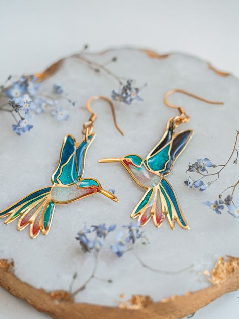 A pair of beautiful blue hummingbird earrings inspired by stained glass compositions.  Each of these bird earrings' holes is filled with different resin tones, creating a beautiful aesthetic, which makes this hummingbird jewelry so magical and colorful. You can choose the handmade earrings' tone from two different tones, gold, and silver. ✦ MEASUREMENT bird pendant: 40 X 20 mm ✦ DETAILS The bird pendants are made of gold or silver-plated stainless steel. The earring hook is hypoallergenic. You c Handpainted Earrings Clay, Magical Jewelry Pendants, Hummingbird Aesthetic, Stain Glass Jewelry, Magical Earrings, Bird Earring, Blue Hummingbird, Hummingbird Jewelry, Stained Glass Earrings