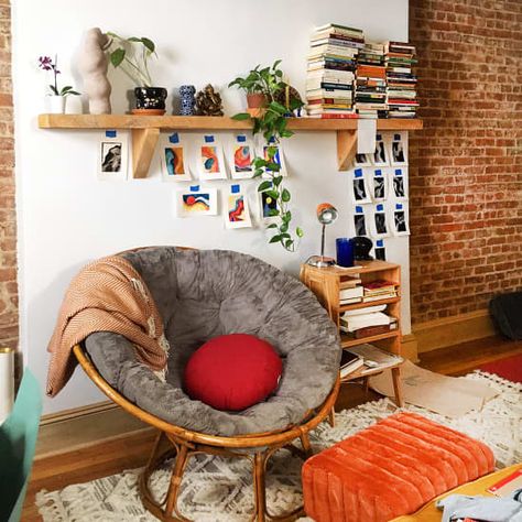 A plush gray papasan chair on a wooden frame, inside an artful bohemian apartment with a brick wall, stacks of books, and small pieces of artwork taped to the wall Small Bohemian Apartment, Papasan Chair Bedroom, Papasan Chair Living Room, Design Intervention, 2020 Bedroom, Bohemian Apartment, Bedroom Corner, Modern Furniture Design, Papasan Chair