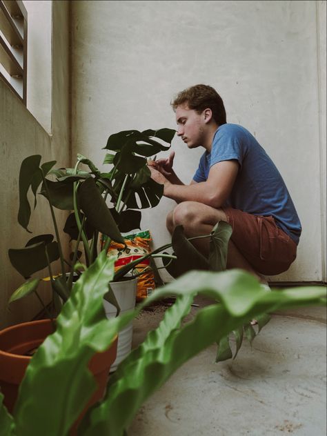 an edited photo of a guy in a blue tshirt and red shorts squatting down touching a small plant in a pot while the camera takes the picture from behind a couple plants taking a candid photo Plant Dad Aesthetic, Plant Parent Aesthetic, Candid Photography Aesthetic, Plant Sunroom, Photo Moodboard, Dad Aesthetic, 60s Aesthetic, Gardening Aesthetic, Lovers Photos