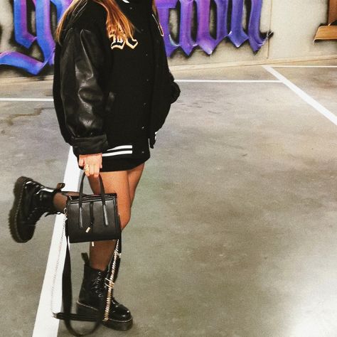 Oversized black varsity jacket with dr Martens Jadon platform boots. Girl posing in an underground parking lot with purple graffiti on the grey walls. She's wearing an oversized varsity jacket in black, half leather with patches stitched on it. She has a small black handbag in her hand with golden chains and she's wearing golden rings and jewelry. Under the jacket she's wearing a black mini skirt with stockings and a black hoodie. Jadon Platform Boots, Black Varsity Jacket, Jadon Boots, Dr Martens Jadon, Dr Martens Boots, Platform Boots, Dr. Martens, Patent Leather, Varsity Jacket