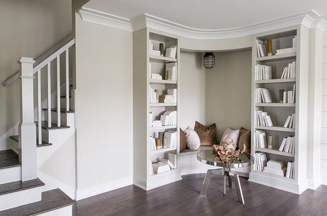 A round chrome coffee table sits in front of a built in corner bench fitted with a gray seat topped with brown and velvet pillows placed against light gray walls lit by a cage pendant light as the bench is flanked by gray built bookcases. Corner Bookcase With Seat, Basement Bench Seating Built Ins, Bedroom Corner Built Ins, Built In Corner Reading Nook, Basement Reading Corner, Built In Reading Corner, Books Corner Ideas, Basement Corner Ideas, Bookcase With Bench