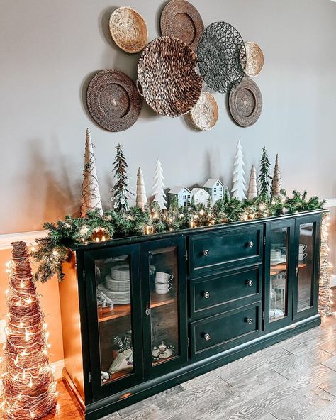 A black farmhouse sideboard with glass doors is set along the wall in this living room. A Christmas village with miniature trees and draping garland is set on top, along with small white lights. Round woven baskets are hung on the wall above, with two twig trees wrapped in white lights are set on either side. Buffet Decor Dining Room, Christmas Sideboard Decor, Buffet Cabinet Decor, Buffet With Glass Doors, Taupe Dining Room, Decorating Sideboards, Decor With Baskets, Sideboard With Glass Doors, Decorating Above Cabinets