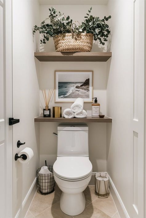 A bathroom with over-the-toilet decor featuring two wooden shelves. The top shelf holds a large woven basket filled with green plants. The lower shelf displays rolled white towels, a scented diffuser, a soap dispenser, and a coastal photograph framed in white. The toilet area also includes a wire basket for toilet paper and a gold wastebasket. The walls are white, creating a clean and serene atmosphere. Small Half Bathroom, Half Bathroom Decor, Toilette Design, Toilet Room Decor, Bathroom Counter Decor, Guest Bathroom Decor, Small Toilet Room, Bathroom Shelf Decor, Over The Toilet