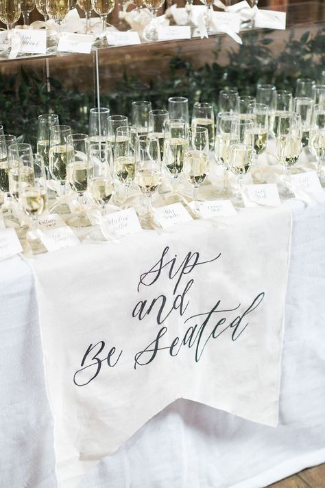 This custom "sip and be seated" matched the adorable placards attached to each glass so as they were seated they could already enjoy themselves. Accented with an acrylic pillar stuffed with greens and it gave us some added height and dimension Sip And Seat Wedding, Take A Sip And Take Your Seat Wedding, Sip And Sit Wedding, Grab A Drink And Take A Seat Sign, Sip And Be Seated Wedding, Welcome Drink Wedding, Champagne Seating Chart, Champagne Seating, Sip And Be Seated