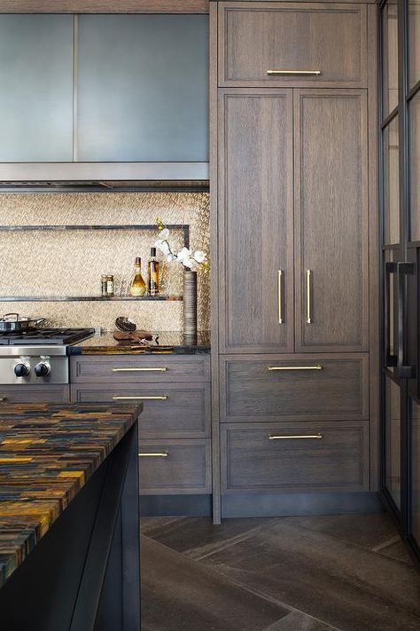 Brown oak kitchen cabinets accented with brass pulls and a gold and black marble countertop are fitted with a stainless steel integrated gas cooktop mounted against gold mosaic backsplash tiles. Brown Oak Kitchen Cabinets, Espresso Kitchen Cabinets, Dark Brown Cabinets, Espresso Kitchen, Brown Kitchen Cabinets, Clean Kitchen Cabinets, Dark Wood Cabinets, Brown Cabinets, Oak Kitchen Cabinets