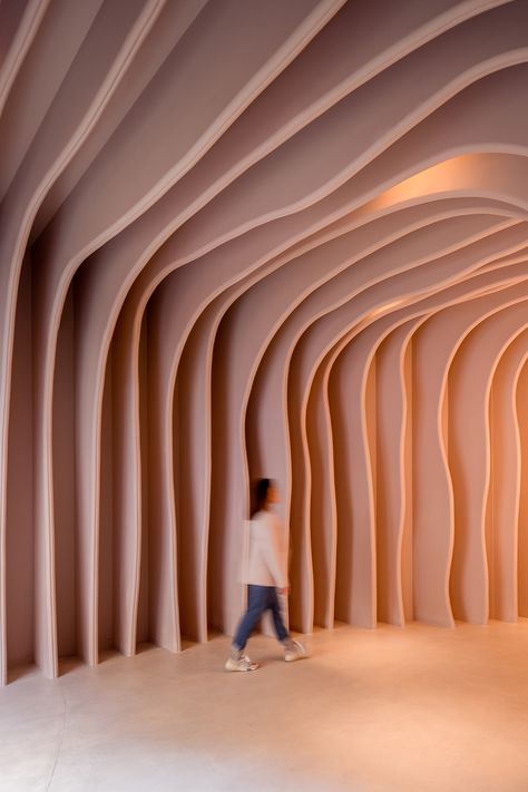 Nude-coloured walls are curved to create a cave-like illusion in a showroom in Barcelona. The walls and ceilings blend into one seamlessly. Showroom Entrance, Wavy Architecture, Curve Wall Design Interiors, Curved Stage Design, Curved Plywood Wall, Curved Exhibition Design, Curved Timber Wall, Acoustic Architecture, Cave Entrance