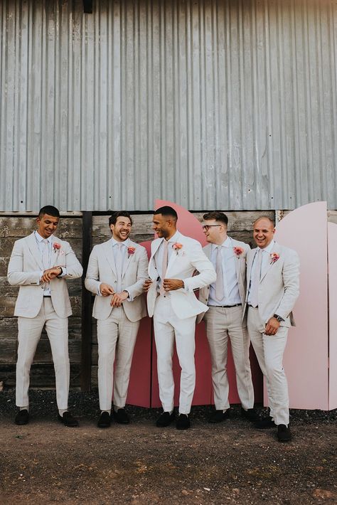 Groom in a white suit and groomsmen in cream suits with pink ties and pink flower buttonholes in front of the pink wedding screen background Groomsmen Colour Scheme, Light Grooms Suit, Bright Wedding Groomsmen, White And Pink Mens Suit, Blush Pink Groomsmen Suit, Groom Blush Wedding, Groomsmen Different Ties, White Suit With Pink Tie, Tan Suit With Pink Tie