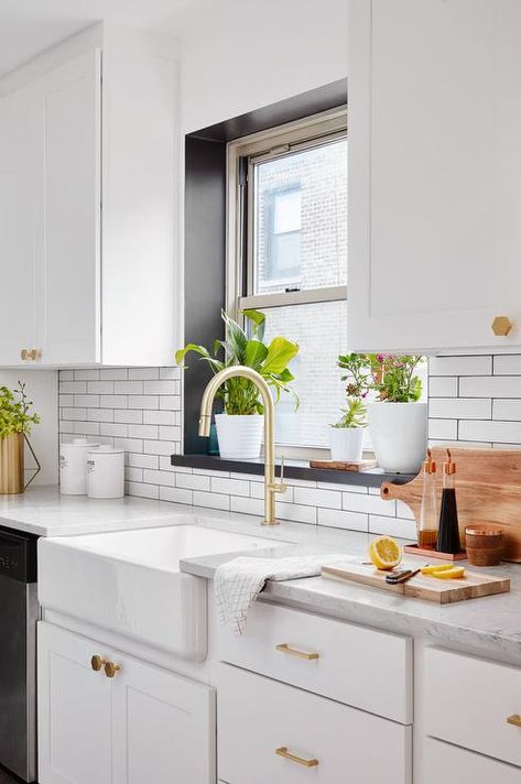 White and gold kitchen features a window framed by a black windowsill positioned above a farmhouse sink with a brushed brass gooseneck faucet fixed to a marble countertop placed against a white subway tile backsplash and on top of white shaker cabinets donning brass hexagon knobs. Window Over Sink, Farmhouse Sink Faucet, Kitchen Window Sill, Farmhouse Backsplash, Easy Home Improvement, White Subway Tile Backsplash, Herringbone Backsplash, Window Seal, Gold Kitchen