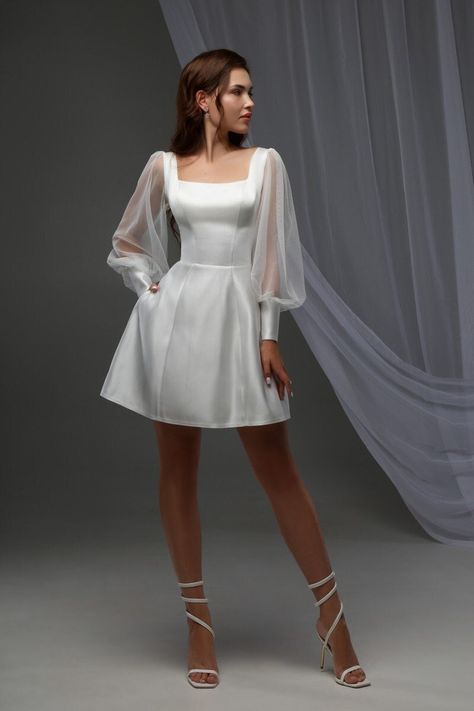 Dive into elegance with our Minimalist White Mini Dress, perfect for any cocktail event or special occasion! Its off-white satin fabric shimmers subtly, making you the center of attention.  - Square neckline & sheer lantern sleeves: A blend of sophistication and charm. - Corset top & natural waistline: Flaunts your figure while the mini length adds a playful twist.  - Pockets & lace-up back: Combine functionality with style. Ideal for summer weddings as a bridal mini dress or an engagement party White Wedding Cocktail Dress, White Bridal Cocktail Dress, Short Balloon Sleeve Wedding Dress, Short Wedding Dress Retro, Cocktail Bridal Dress, Short Reception Dress For Bride Fun, Mini Wedding Dress With Sleeves, Skater Wedding Dress, Short White Vintage Dress